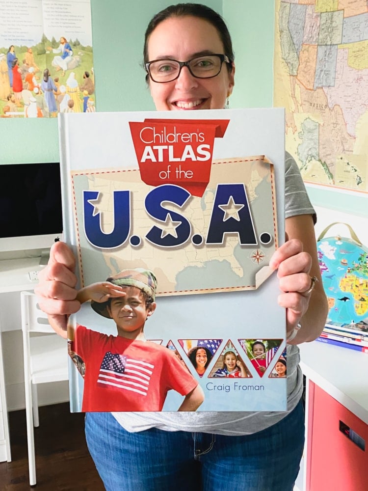 a woman holding out in front of her a large children's atlas of the usa book