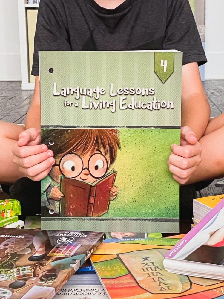 a boy holding a book called language lessons for a living education 4 with more books all around him