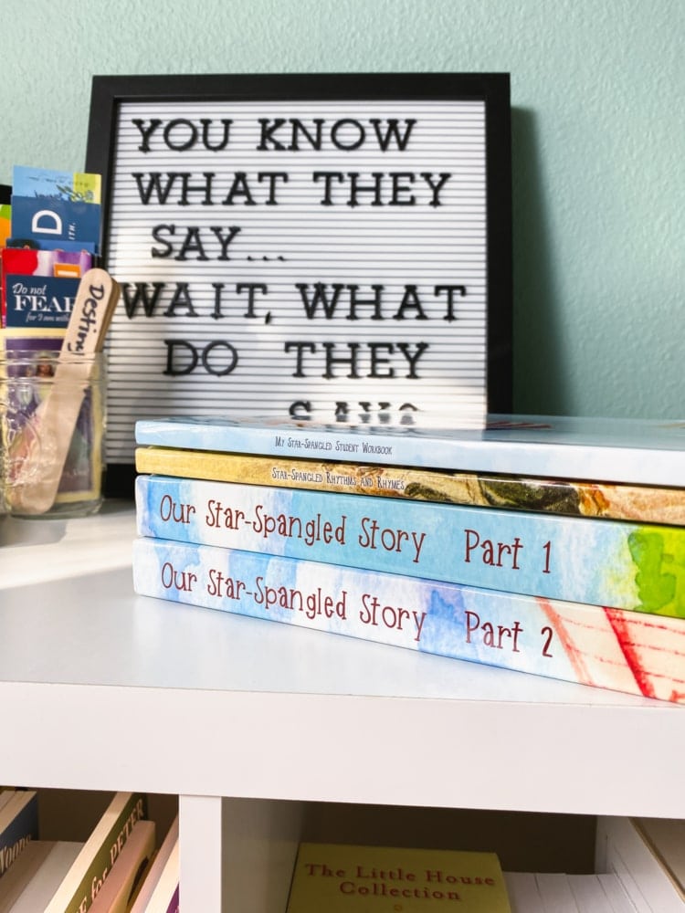 a shelf with 4 books from our star spangled story homeschool history curriculum and some school supplies in background