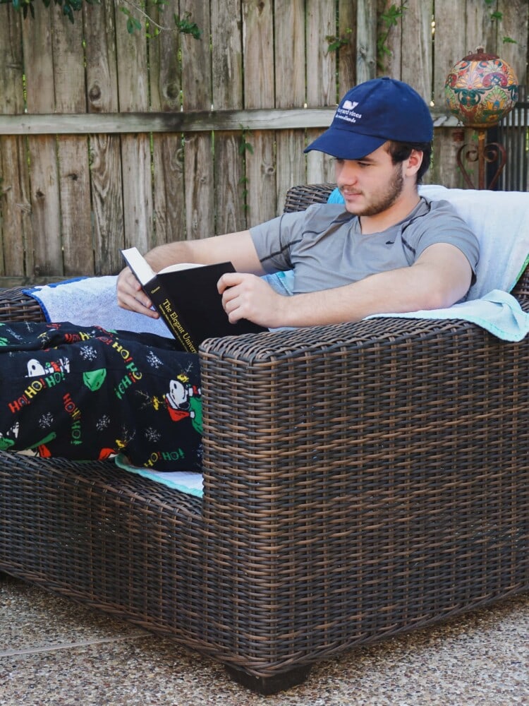 highschool boy reading a book for homeschool while sitting in a lounge chair outside on the patio