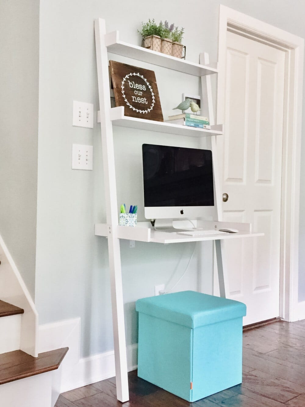 a small ladder desk leaning against the wall with a small popup stool underneath and a desktop computer and decor sitting on the upper shelves