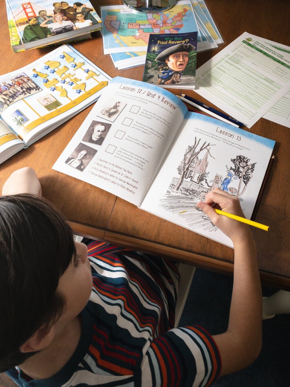 a young boy sitting at a desk with many books, worksheets, and coloring pencils laid out on top of the desk, coloring in a coloring book that is part of the Our Star-Spangled Story curriculum